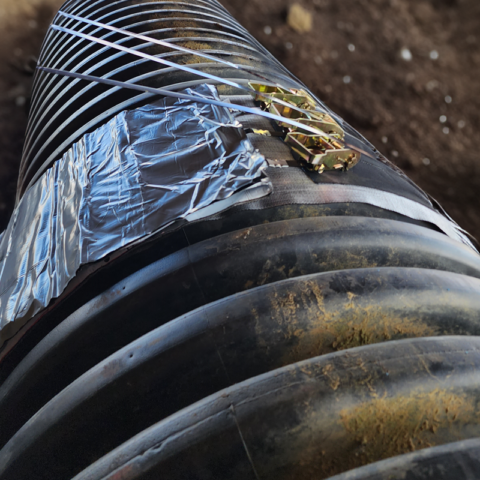 Black corrugated plastic pipe laying in a dark brown ditch with a mastic like type wrapped around the joining ends
